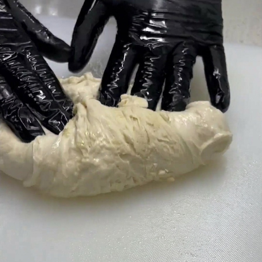 Une personne portant des gants noirs qui étire et replie un morceau de pâte pâle et humide sur une surface blanche.