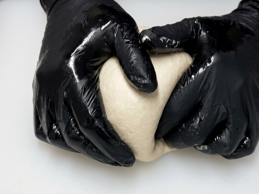 Des mains avec des gants noirs façonnant une boule de pâte à pizza ronde et lisse en la faisant glisser sur une planche blanche.