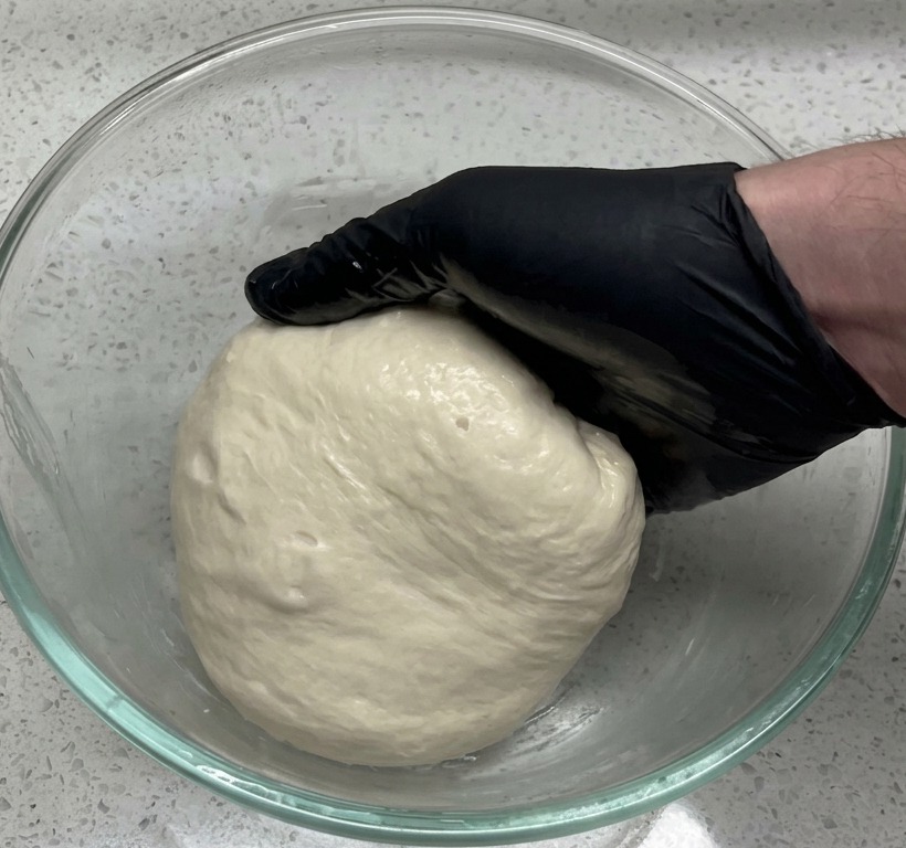Une main gantée de noir plaçant une boule de pâte lisse dans un grand saladier en verre transparent posé sur un plan de travail de style granit.