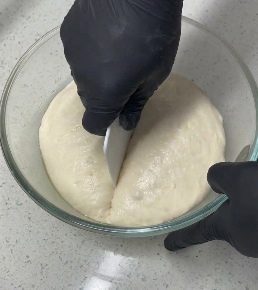 Une personne avec des gants noirs utilise un coupe-pâte blanc pour diviser en deux une grosse masse de pâte aérienne dans un saladier en verre.