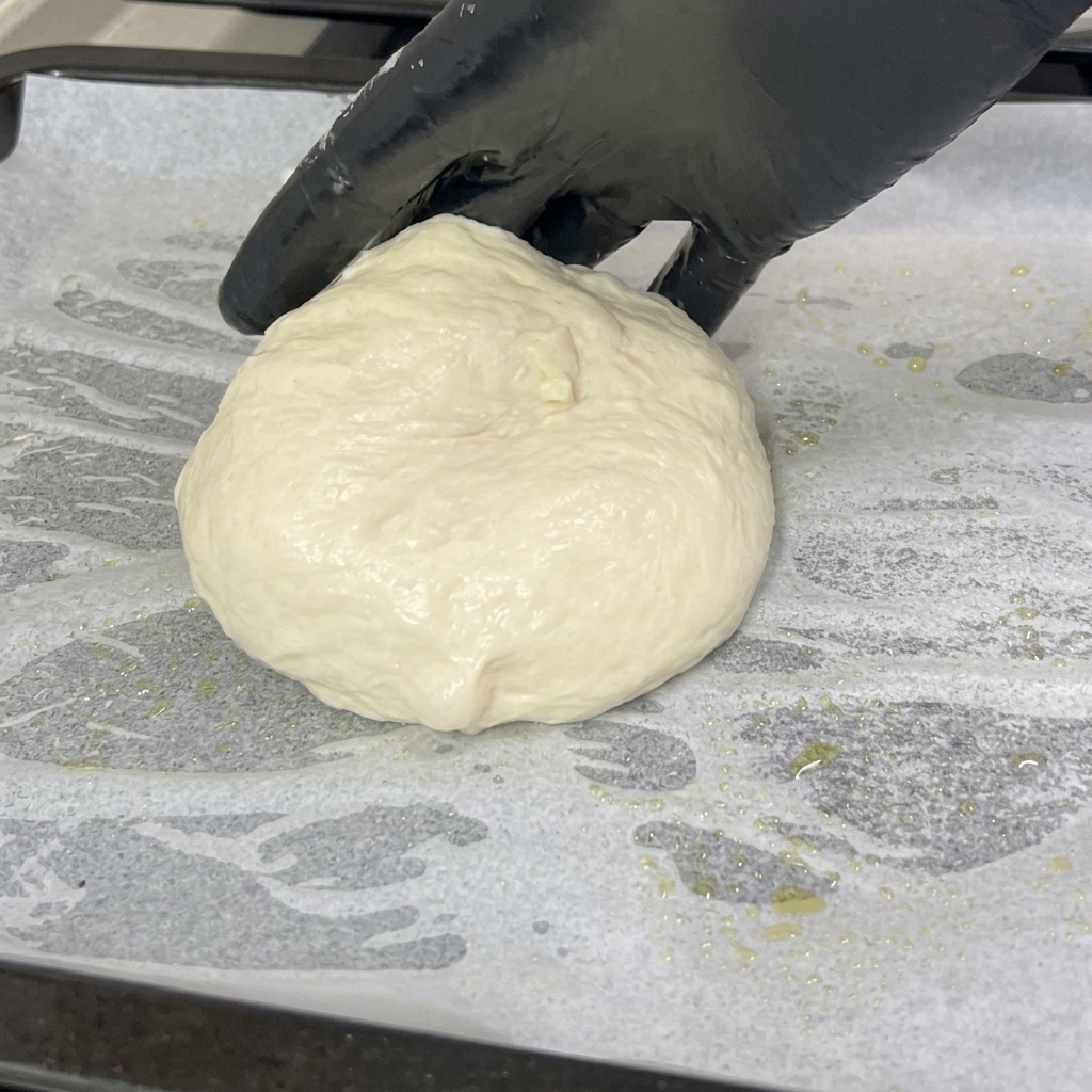 Une main gantée déposant une boule de pâte à pizza souple et arrondie sur une plaque recouverte de papier sulfurisé blanc.