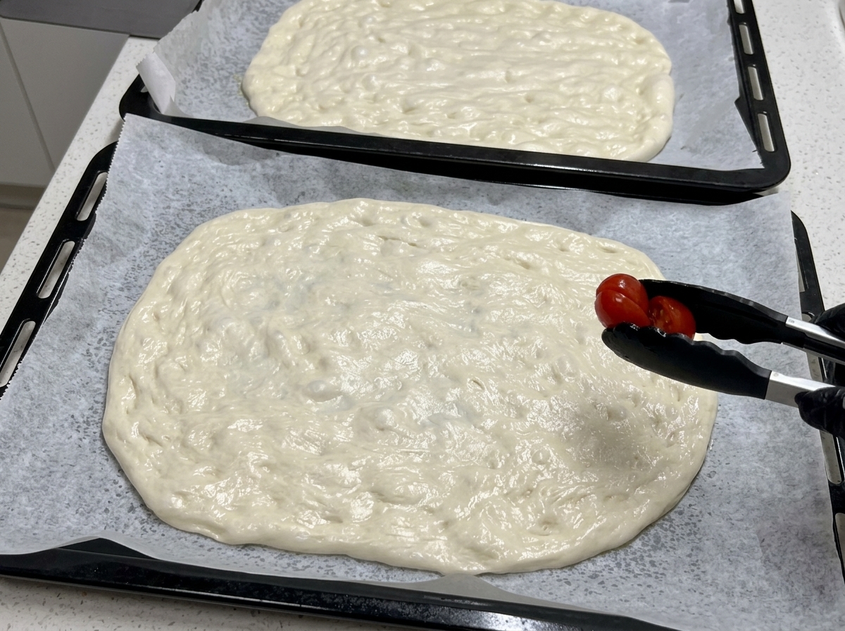 À laide dune pince de cuisine noire, une demi-tomate cerise est déposée sur une grande abaisse de pâte à pizza crue.