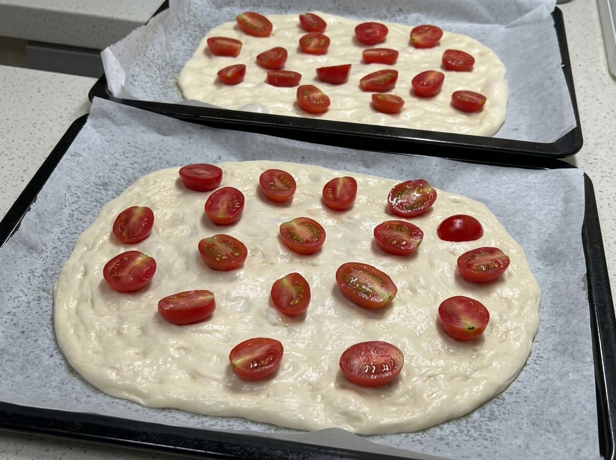 Deux plaques de cuisson montrant la pâte à pizza étalée et garnie de nombreuses demi-tomates cerises, prêtes pour lassaisonnement.
