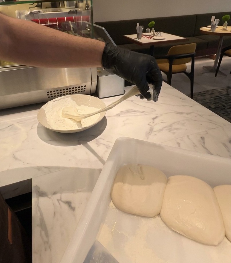 Une main gantée soulevant un pâton de pizza élastique et fermenté hors dun bol de farine blanche.