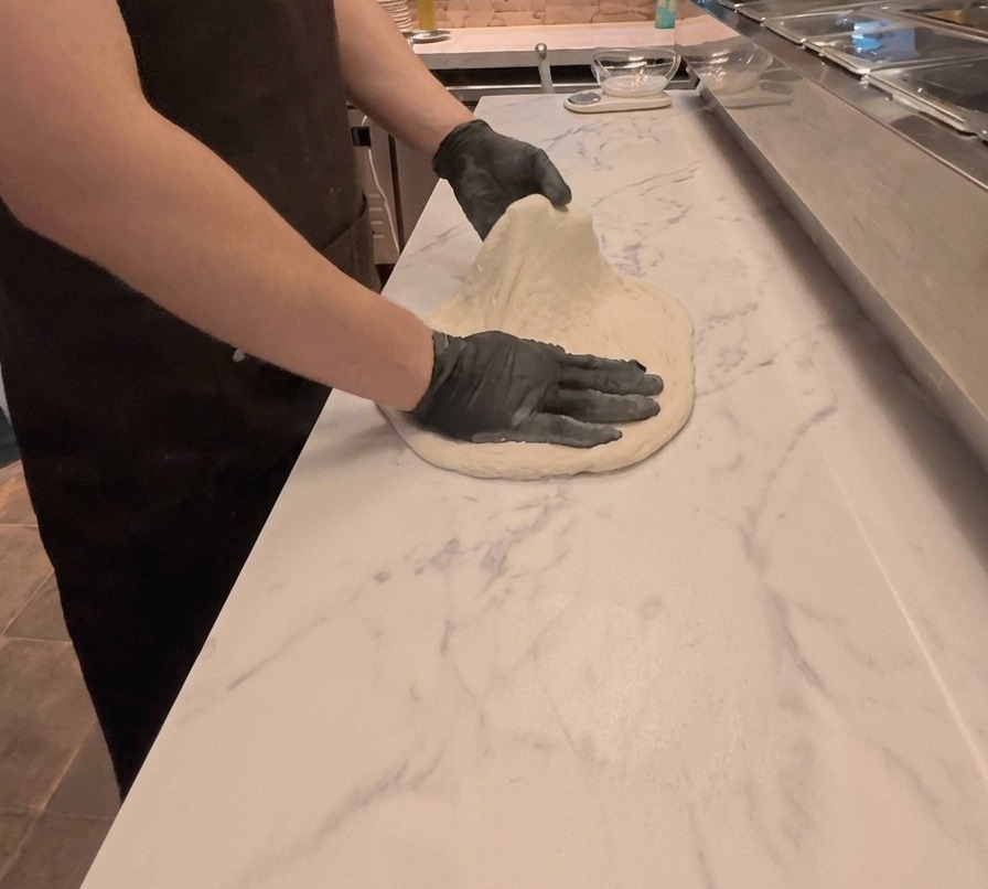 Un cuisinier étirant et aplatissant à deux mains la pâte à pizza sur un plan de travail en marbre blanc.