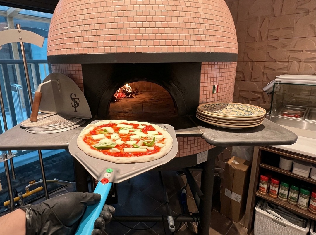 Une pizza crue garnie de sauce tomate et de mozzarella posée sur une pelle métallique à lentrée dun four à bois en briques roses.