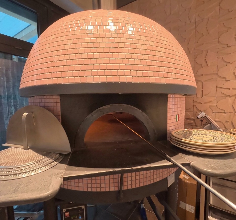 Lextérieur dun four à pizza en dôme avec des briques roses, laissant entrevoir une pizza en train de cuire près des braises incandescentes.