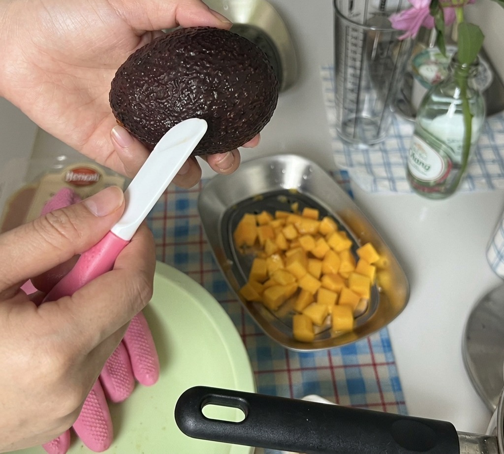 Mains coupant une moitié davocat pelée en petits cubes au-dessus dun plateau contenant déjà de la mangue en dés.