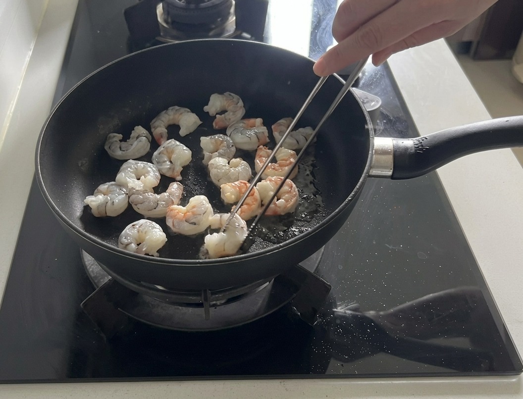 Crevettes fraîches sautées dans une poêle noire avec de lhuile dolive, certaines devenant roses et opaques.