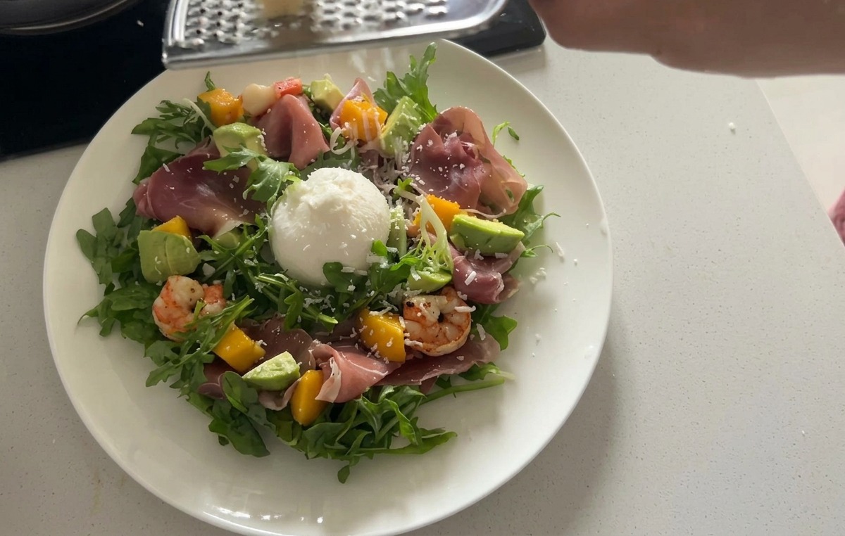 Une salade de roquette dressée dans une assiette, surmontée dune grosse boule de burrata blanche et de fines tranches de prosciutto chiffonnées.