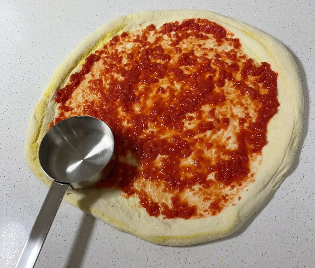 Une louche en métal étalant de la sauce tomate bien rouge en mouvements circulaires sur une pâte à pizza crue.