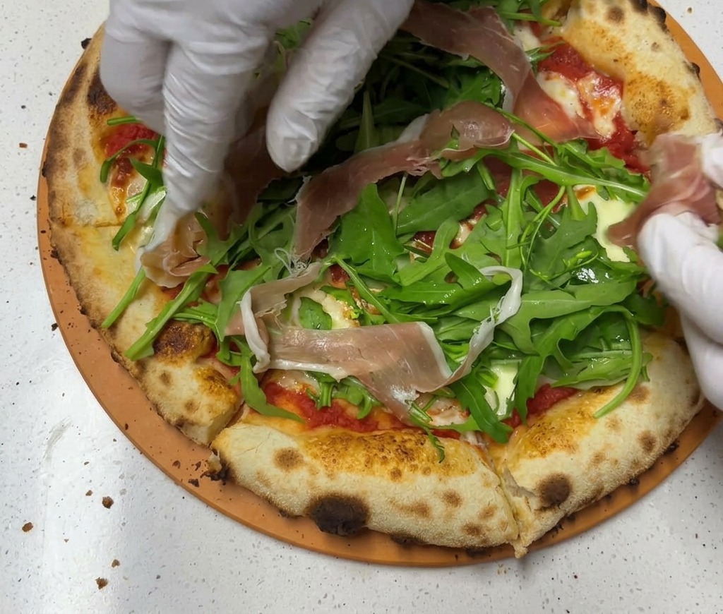 Des mains déposant de fines tranches de jambon de Parme rosé sur le lit de roquette dune pizza cuite.