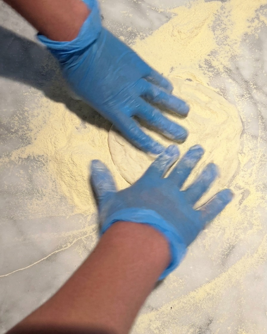 Mains portant des gants bleus pressant une boule de pâte à pizza sur un lit de semoule de blé sur un plan de travail en marbre.