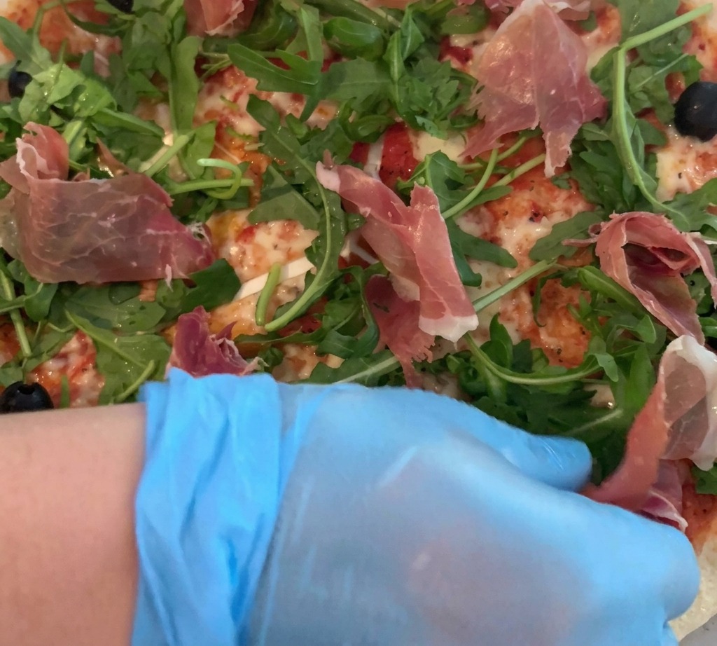 Une main gantée déposant délicatement de fines tranches de jambon cru sur une pizza garnie de roquette fraîche.
