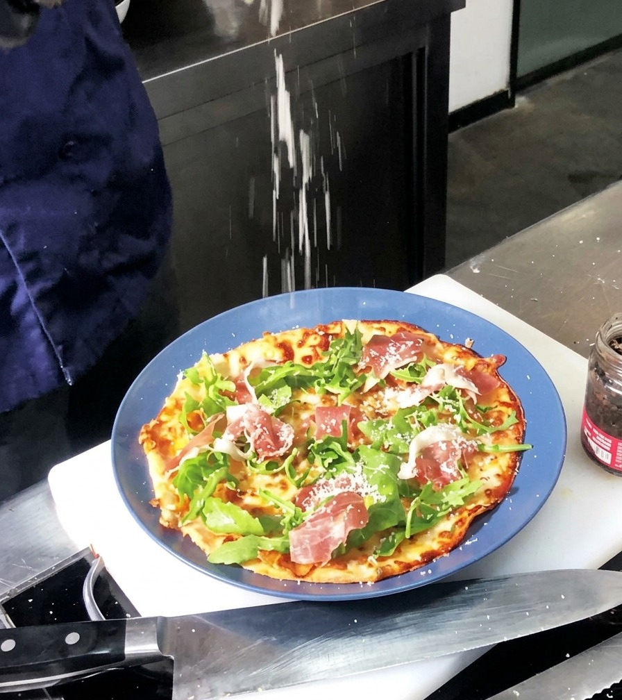 Du parmesan fin en poudre saupoudré sur une pizza cuite garnie de roquette verte et de tranches de jambon rouge.