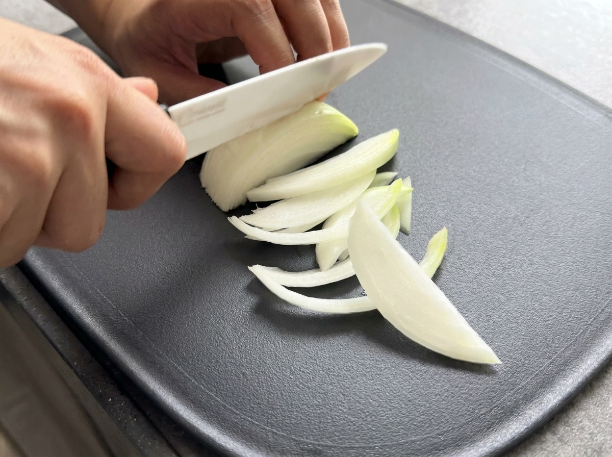 Gros plan dun couteau tranchant un demi-oignon en fines lanières uniformes sur une surface sombre.