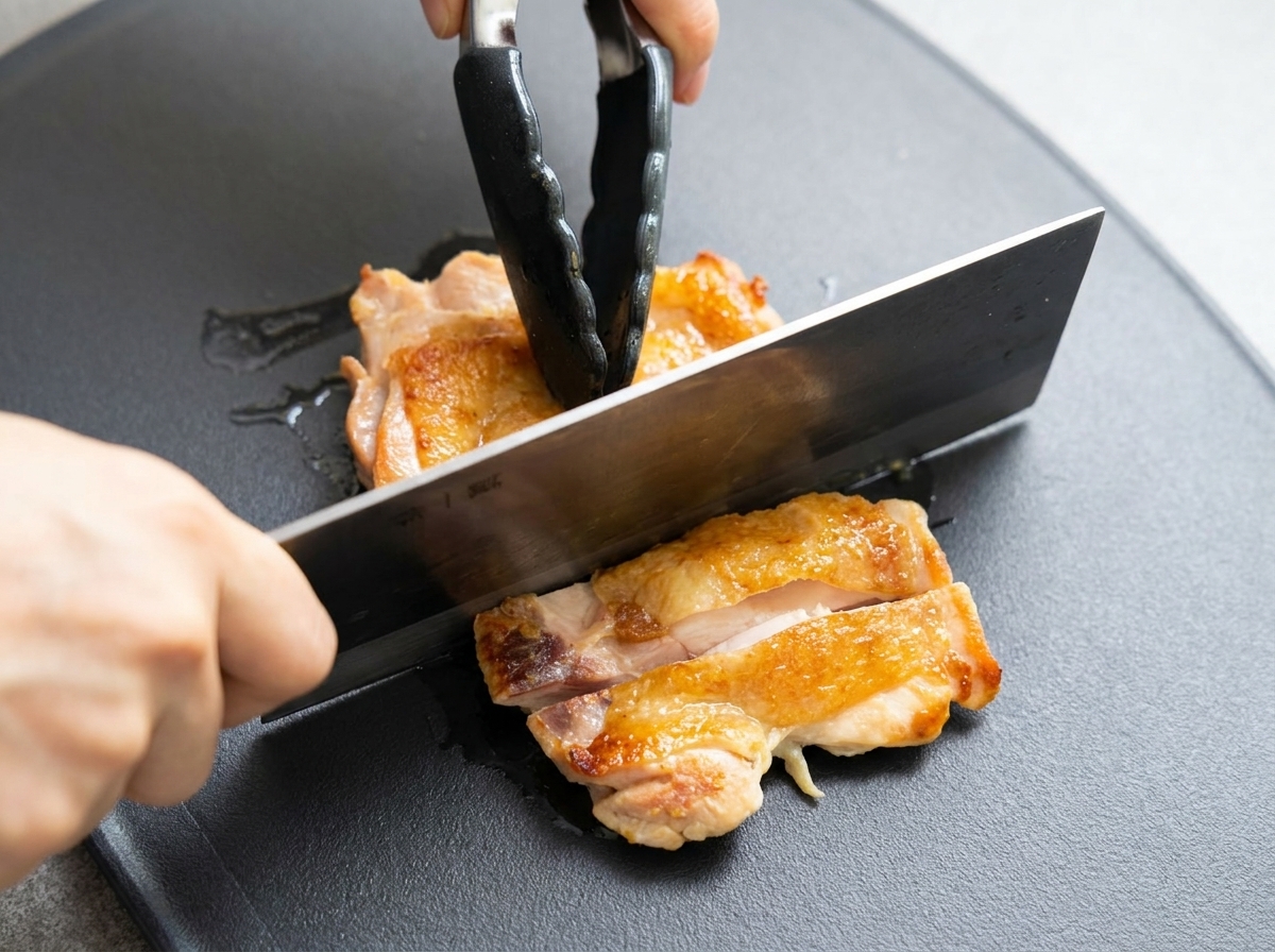 Cuisse de poulet dorée en train dêtre tranchée en morceaux sur une planche à découper sombre avec un couperet.