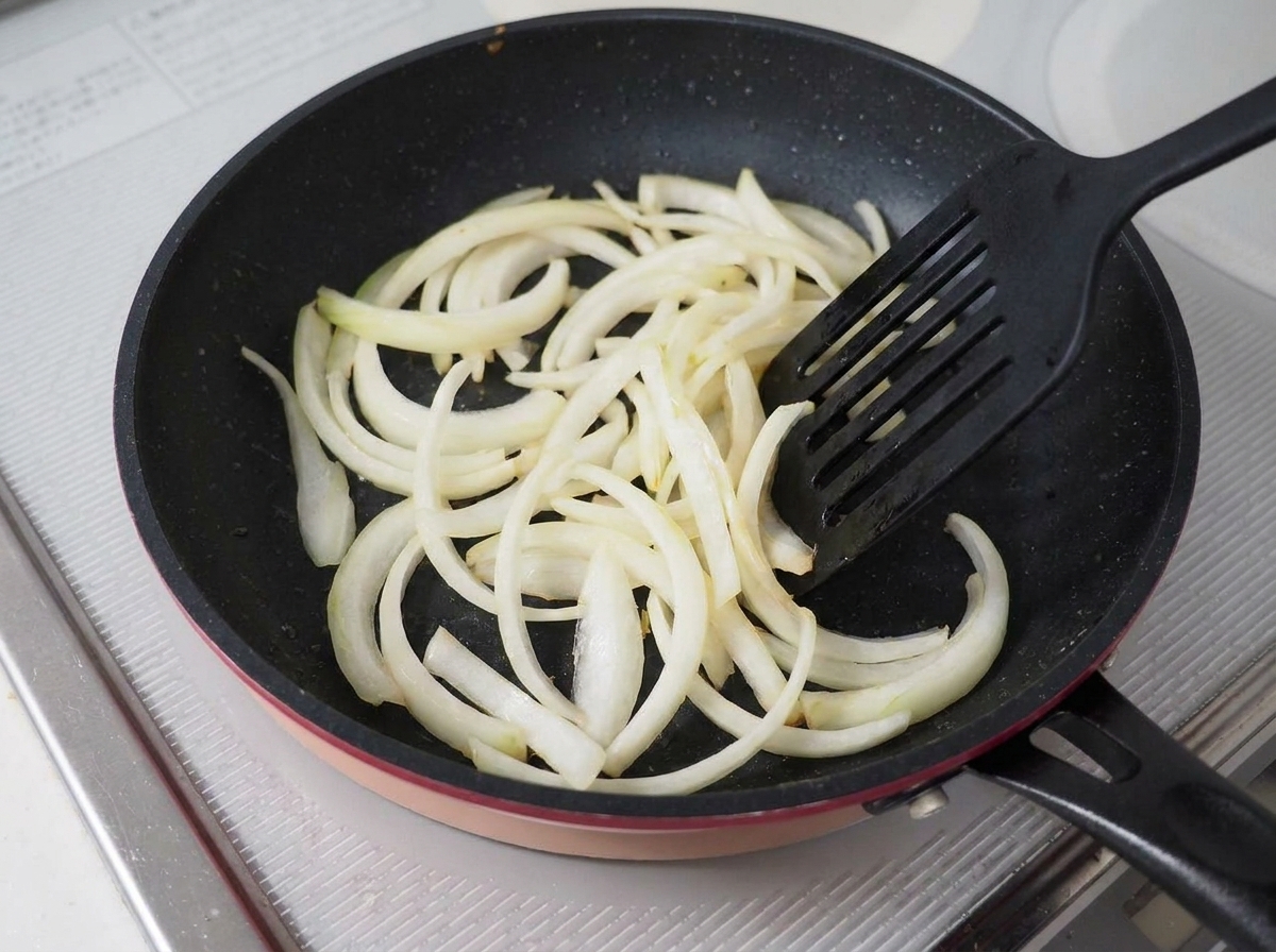 Oignons émincés en train de revenir dans une poêle antiadhésive sombre avec une spatule noire.