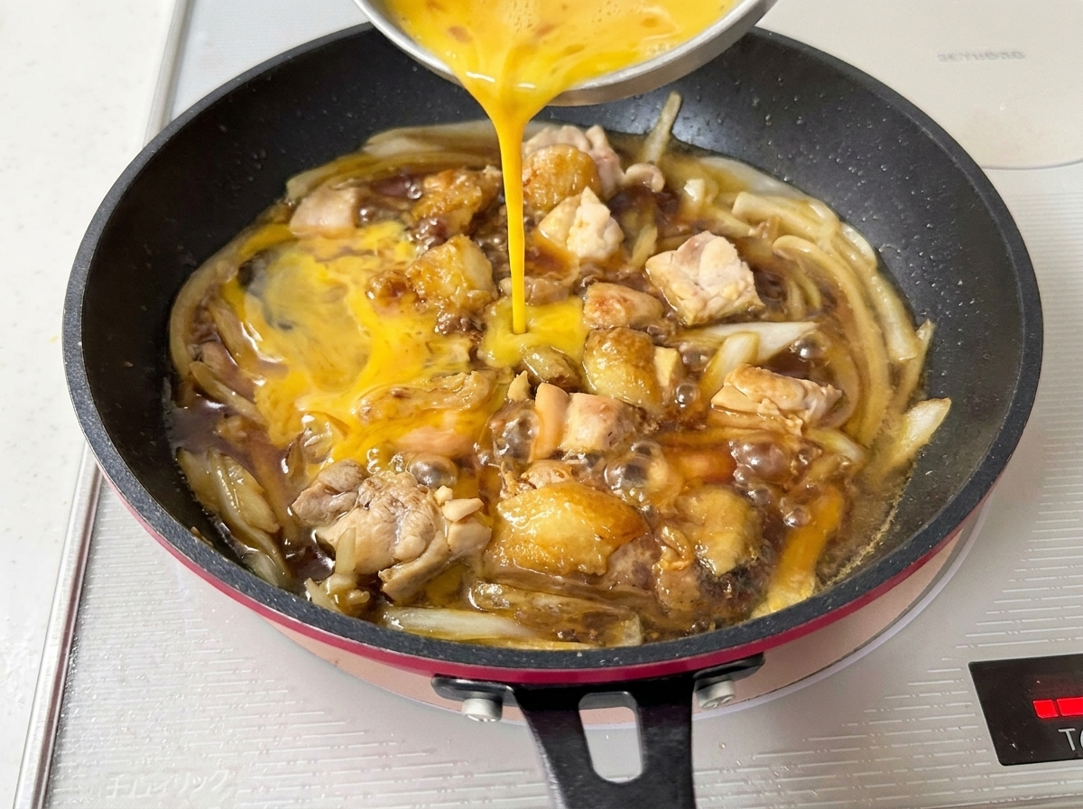 Œufs battus versés depuis un bol dans la poêle, nappant le poulet et les oignons mijotants.