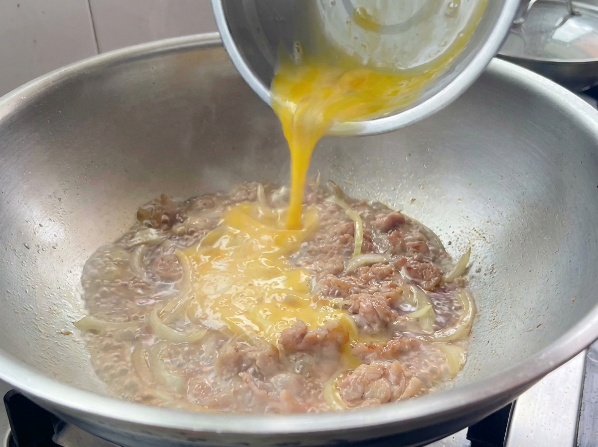 Mélange dœufs battus versé sur le porc et les oignons dans une poêle.