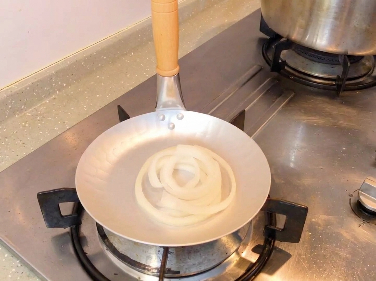 Mélange de sauce soja noire versé à la cuillère dans une poêle contenant des rondelles doignon blanc cru.