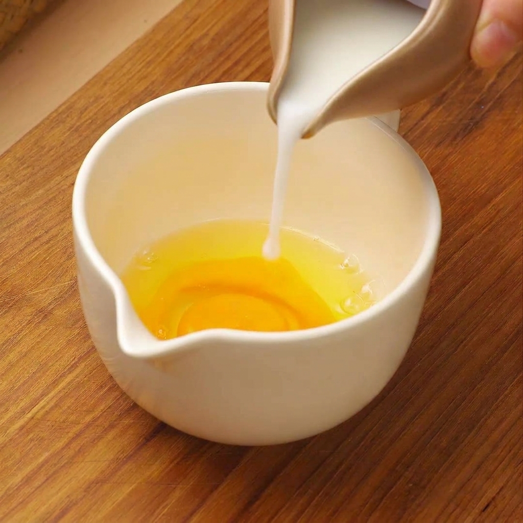 Du lait blanc versé depuis un petit pichet dans un verre doseur contenant des œufs crus cassés.