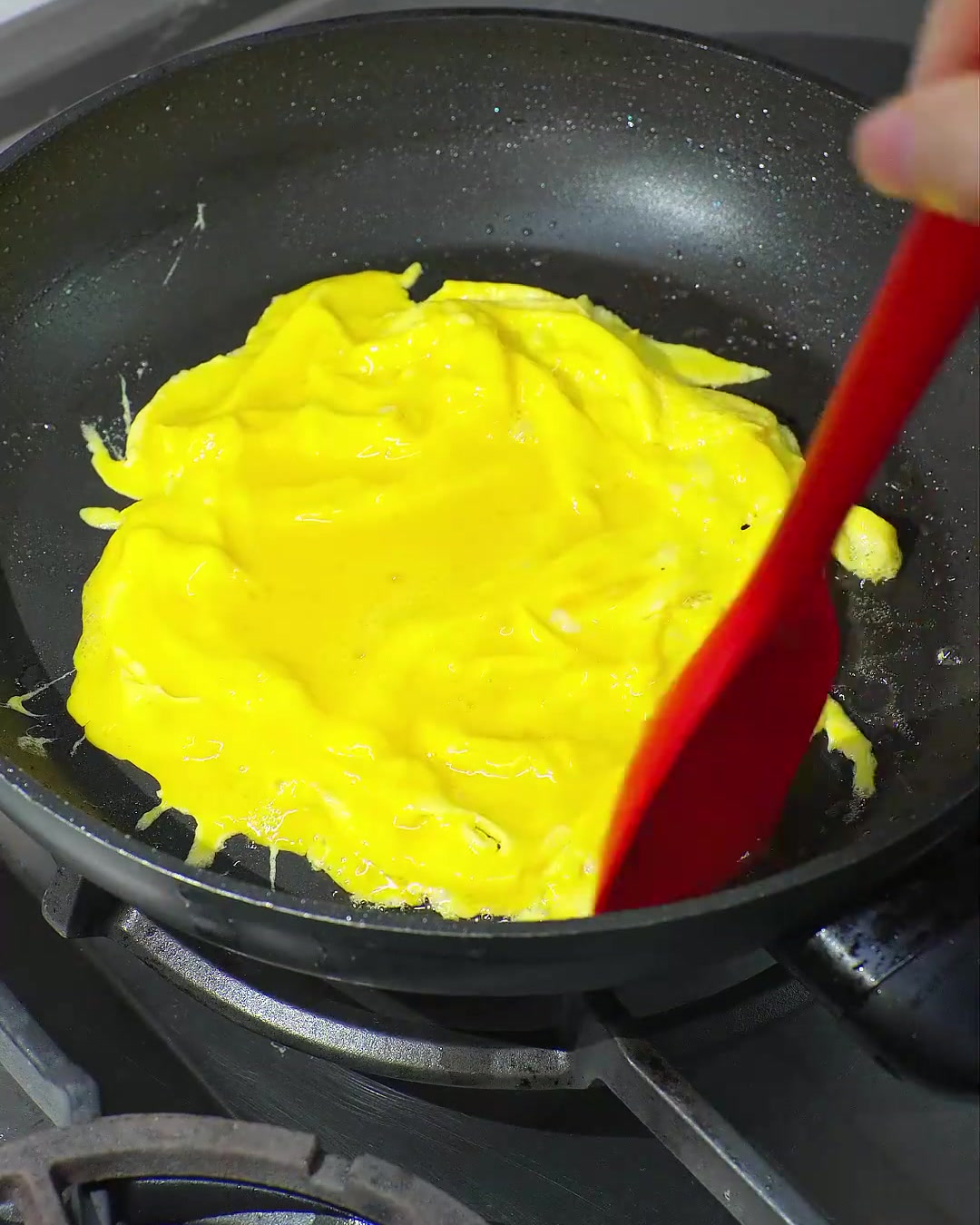 Une spatule rouge repliant un œuf brouillé jaune vif partiellement cuit dans une poêle noire.