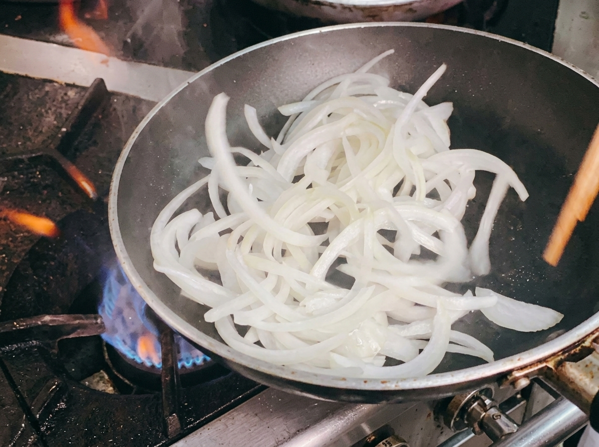 Des oignons blancs émincés sautés dans une poêle à laide de baguettes en bois.