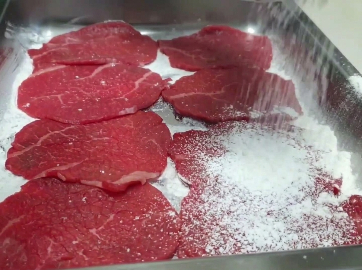 Main saupoudrant de la farine blanche sur des tranches de bœuf crues disposées sur une plaque.