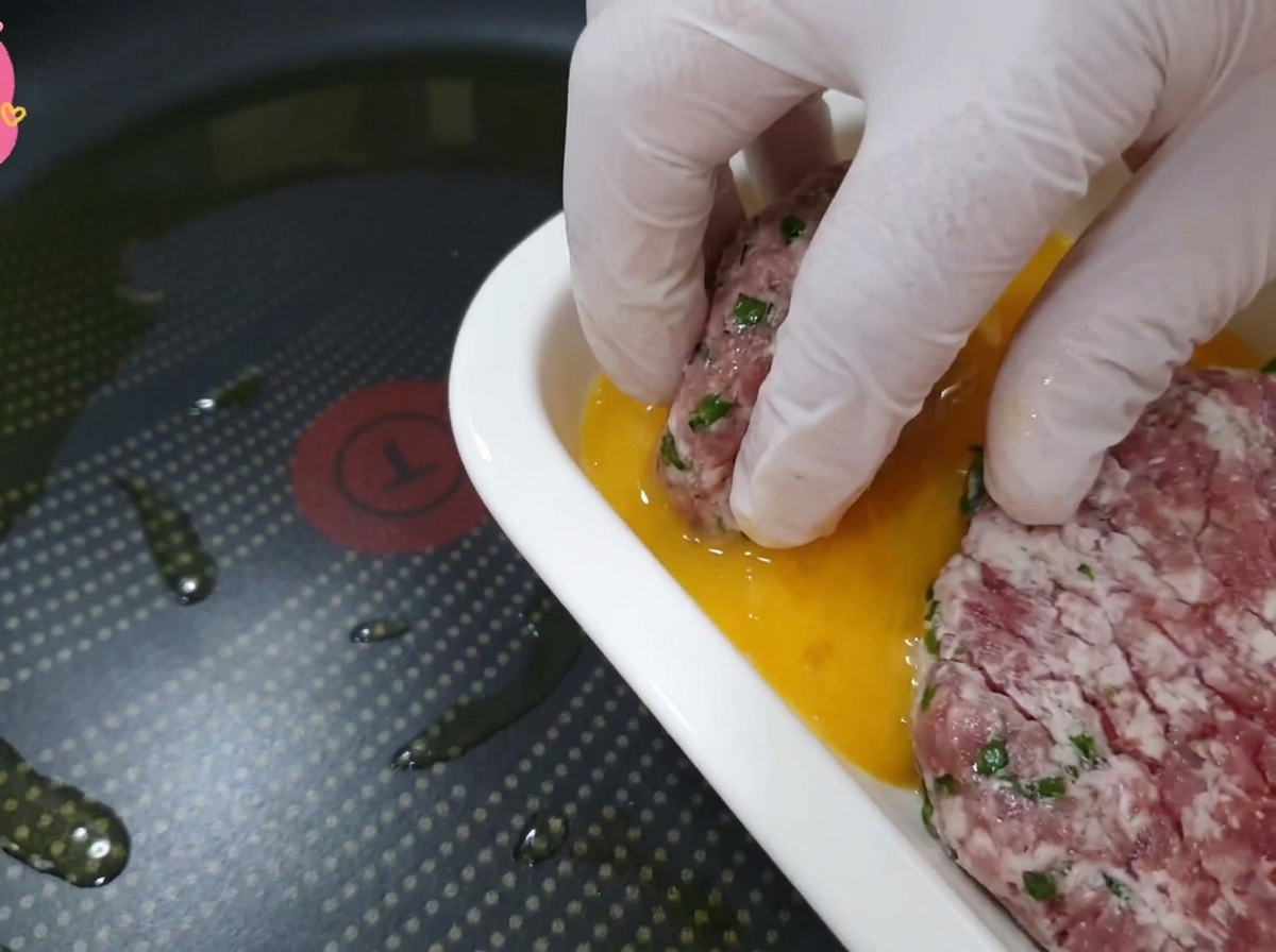Une main gantée trempant un morceau de bœuf et de feuille de perilla dans un plat rempli dœufs battus dorés.
