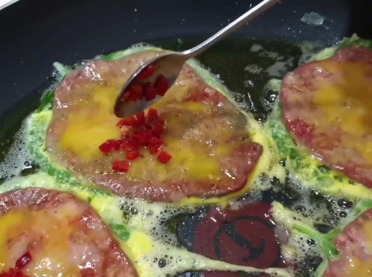 Gros plan sur une cuillère ajoutant des piments verts et rouges coupés en dés sur le dessus des galettes de bœuf en train de cuire dans une poêle.