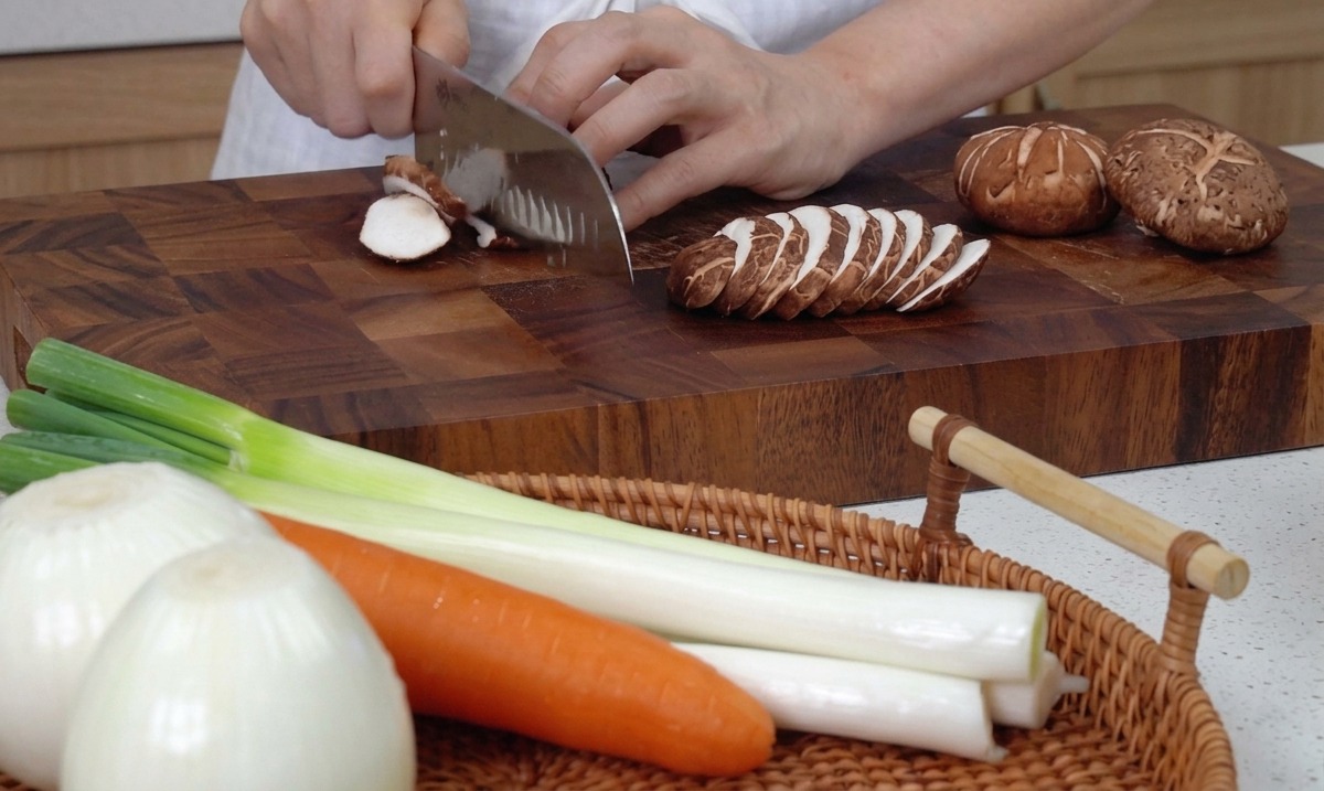 Mains utilisant un grand couteau de cuisine pour trancher des champignons shiitake frais sur une planche en bois.