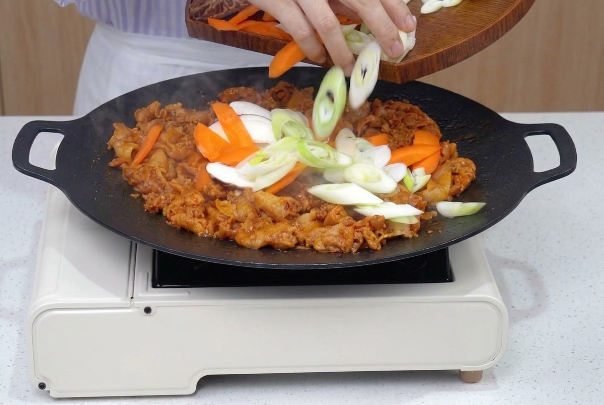 Une poêle remplie de porc doré rejointe par des tranches fraîches de carottes, oignons et champignons.