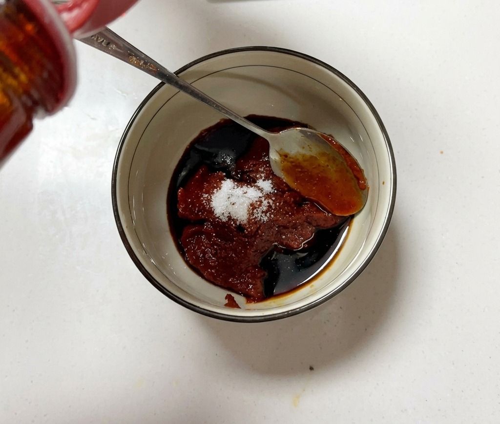Un petit bol rempli de pâte de piment coréenne, de sucre et de sauce soja en cours de mélange avec une cuillère en métal.