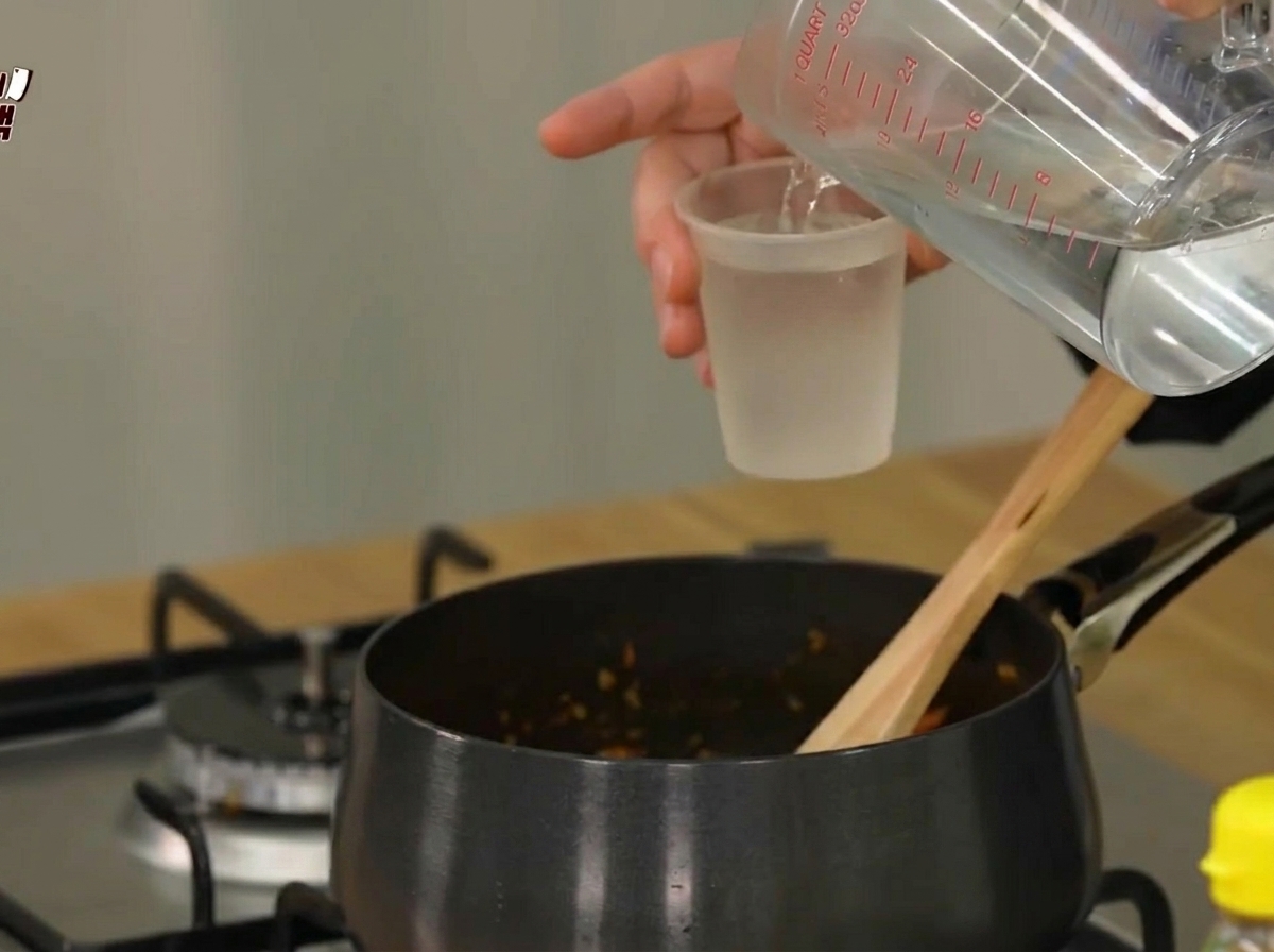 De leau versée dans une casserole noire contenant les ingrédients sautés.