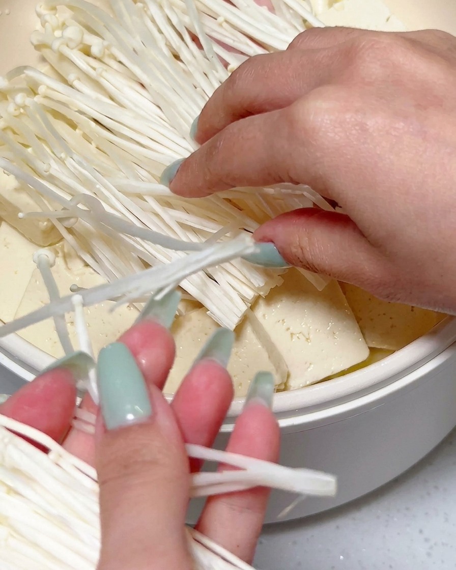 Mains déposant une touffe de champignons enoki frais sur le tofu et les autres ingrédients dans la poêle.