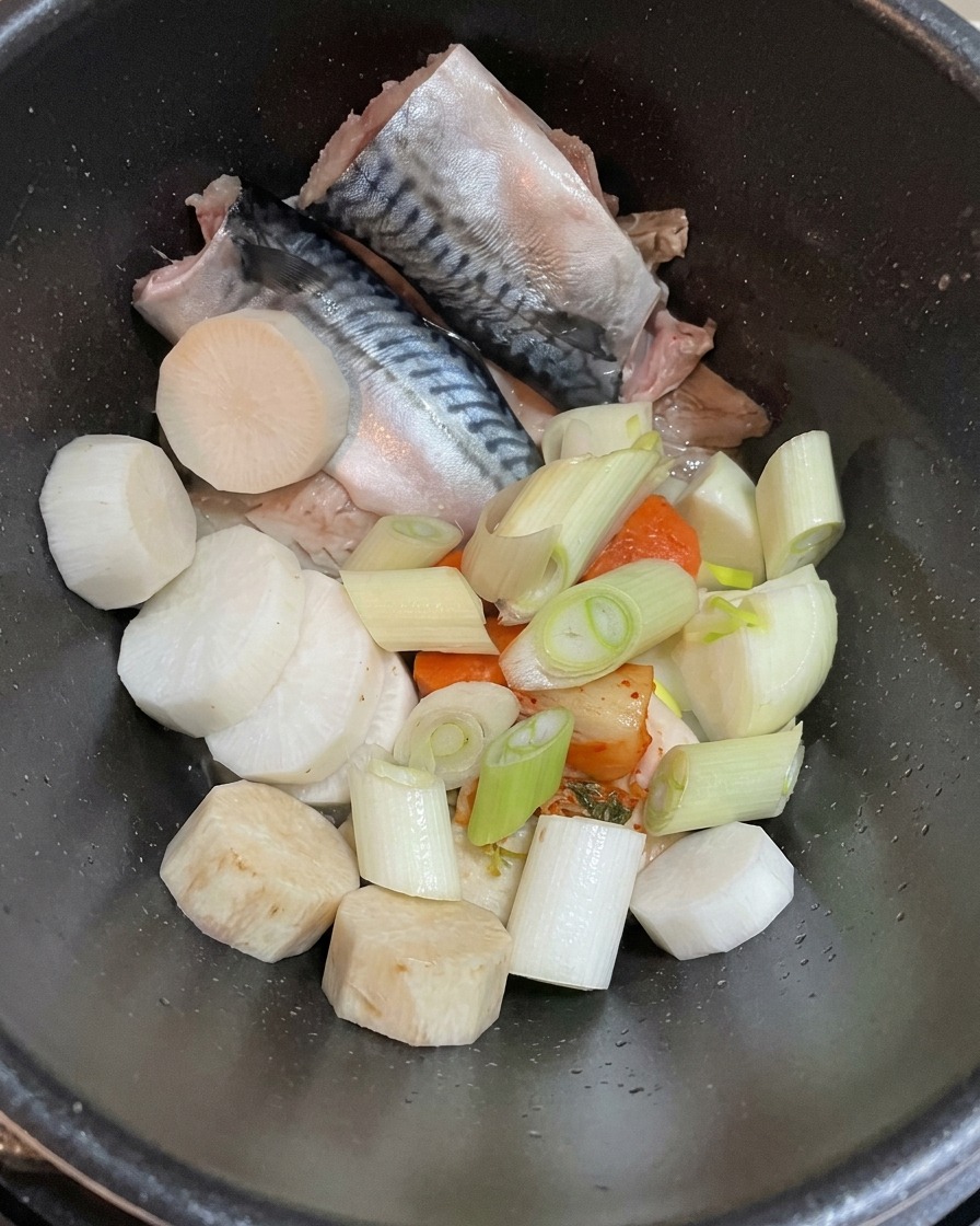 Vue de dessus dune casserole sombre contenant des filets de maquereau crus, des morceaux de radis blanc et des oignons verts émincés.