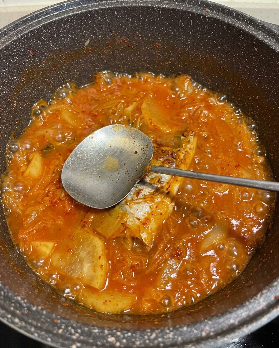 Un ragoût rouge bouillonnant de maquereau et de légumes dans une casserole, avec une cuillère en métal soulevant un morceau de poisson.