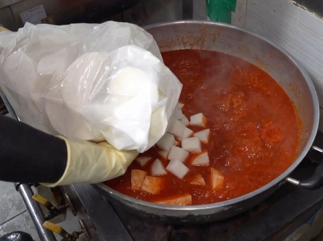 Des morceaux de radis ajoutés à une grande casserole remplie de liquide épicé rouge bouillant.