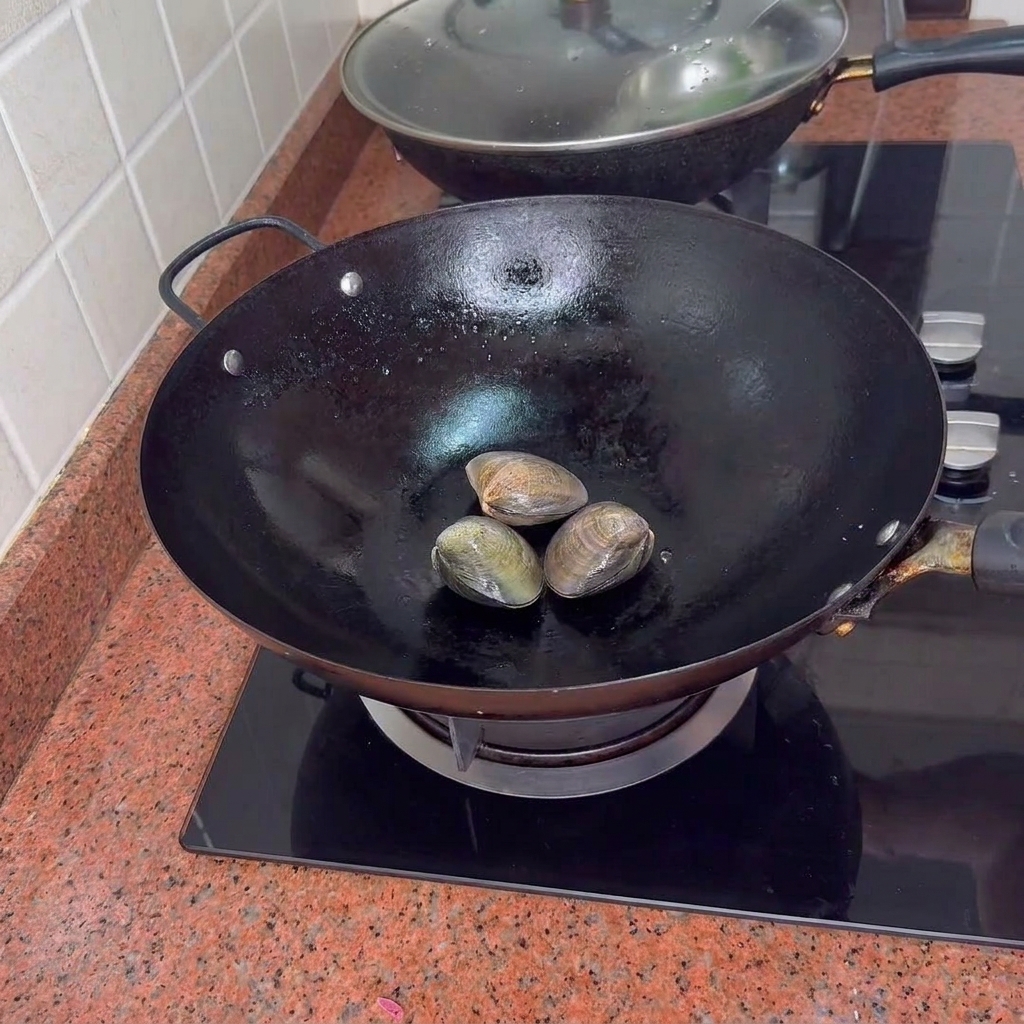 Trois palourdes fermées dans un wok noir sur une plaque de cuisson.