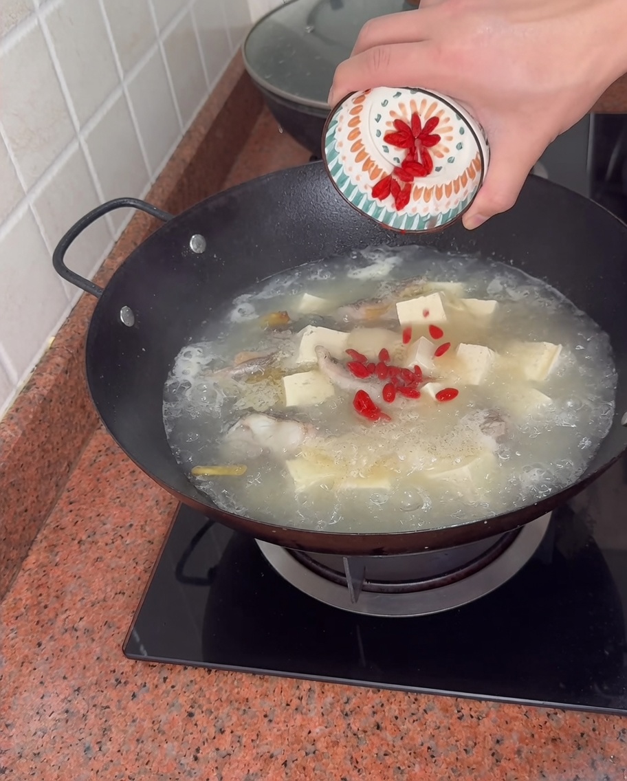 Verser des baies de goji rouges depuis un petit bol à motifs dans un wok bouillonnant de soupe de poisson et tofu.