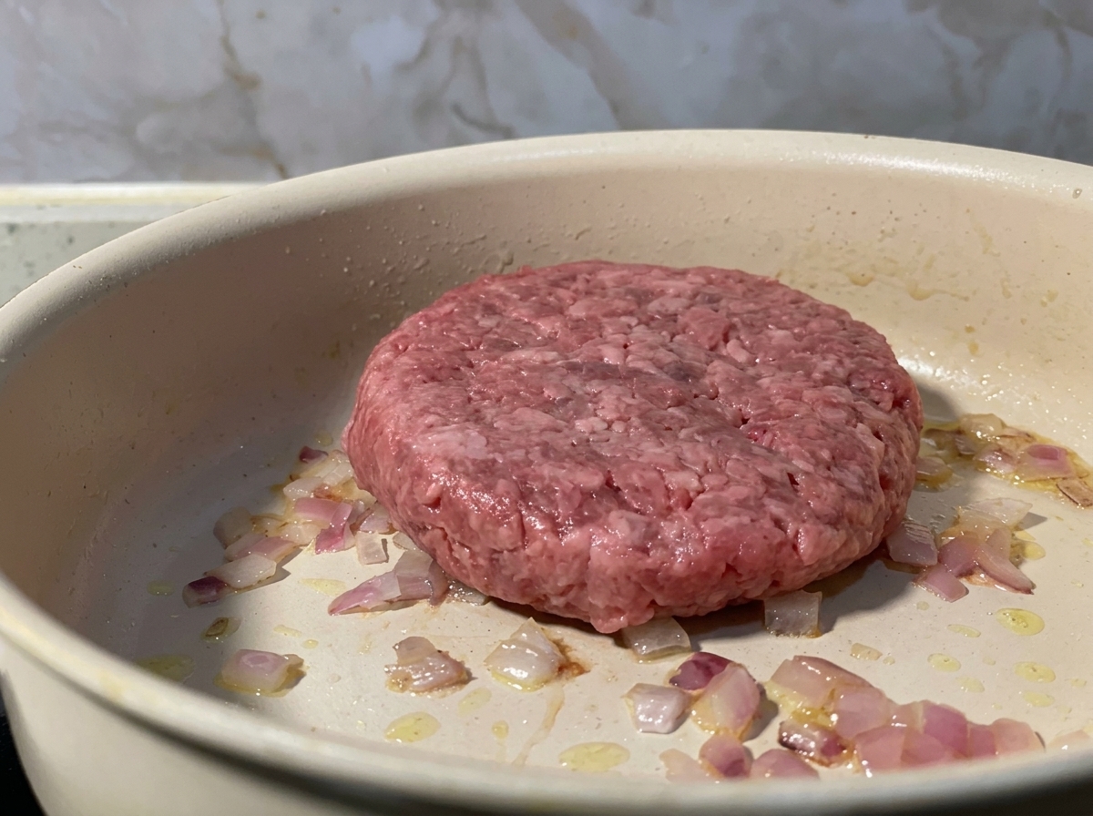 Un steak de bœuf haché cru placé dans une poêle avec des oignons rouges revenus.