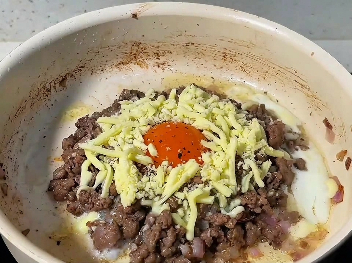 Fromage râpé couvrant le bœuf et lœuf dans une poêle, le tout couvert dun couvercle.