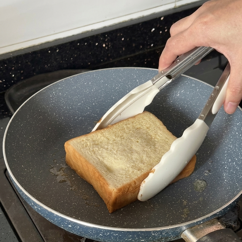 Une tranche de pain épaisse toastée dans une poêle avec des pinces blanches.