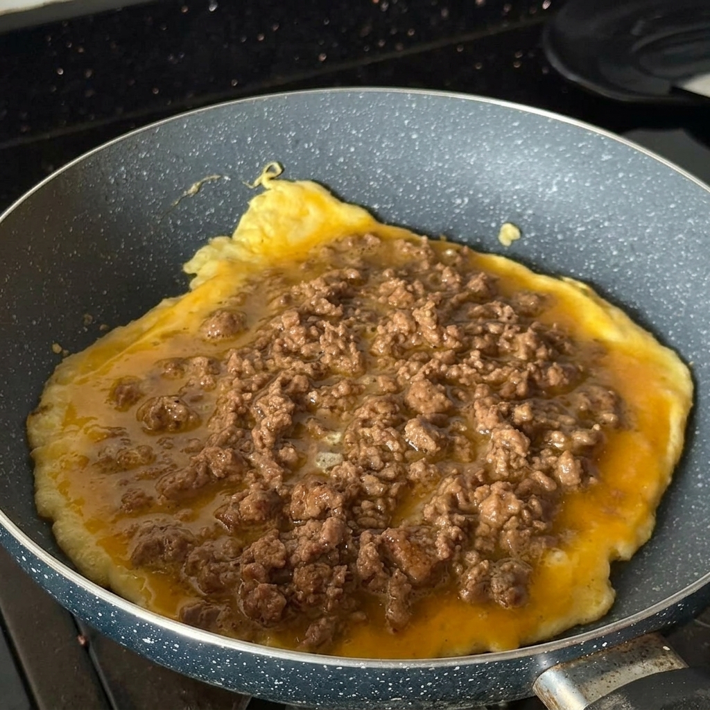 Une omelette ronde cuisant dans une poêle mouchetée, généreusement recouverte de bœuf haché cuit et assaisonné.