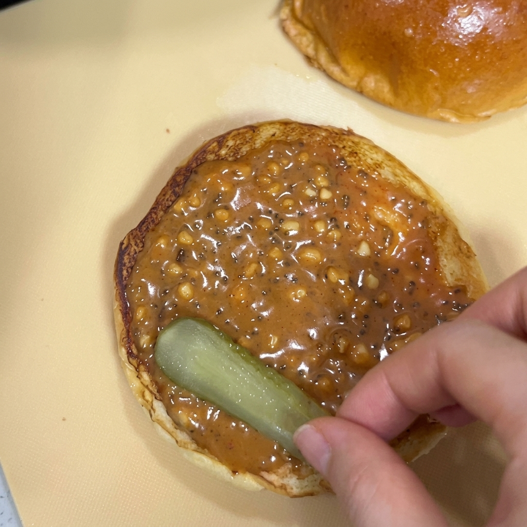 Une main déposant une tranche de cornichon sur le pain couvert de sauce cacahuète.