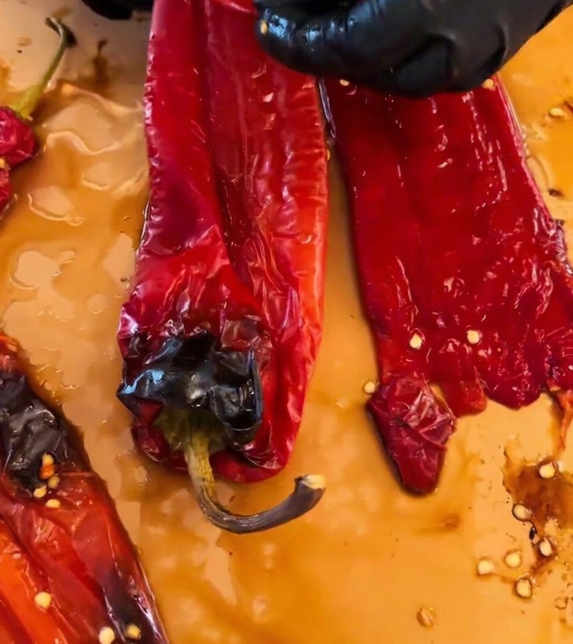 Mains portant des gants noirs pelant la peau brûlée de piments rouges rôtis sur une surface de travail.