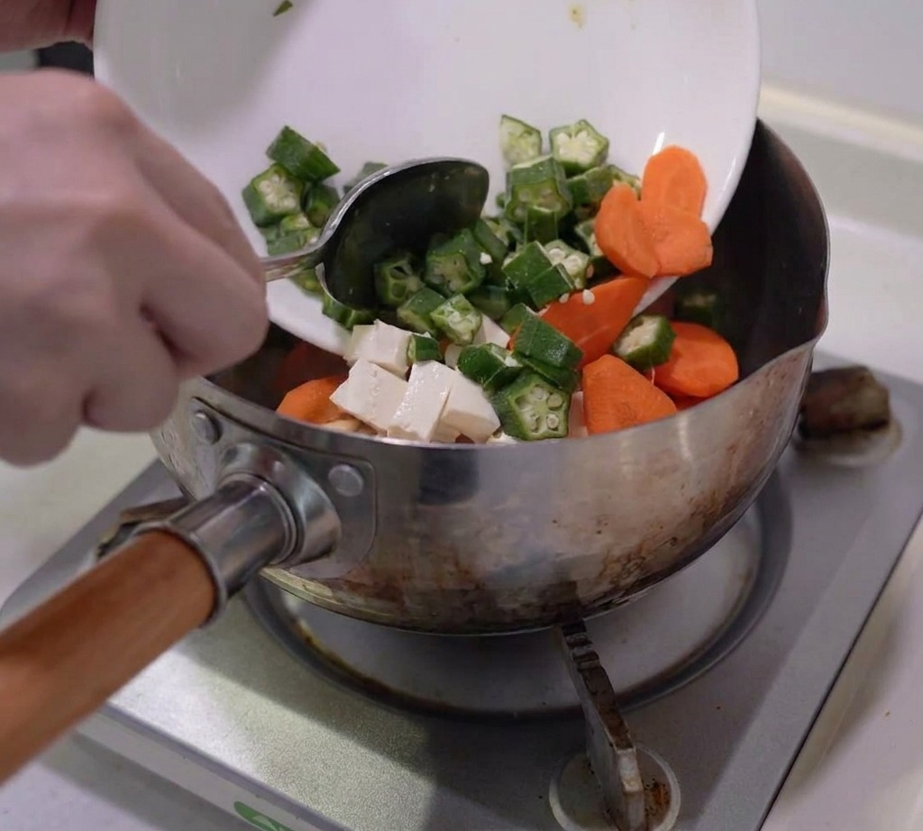 Gombo tranché, morceaux de carotte, champignons et tofu déposés dans une casserole en inox.