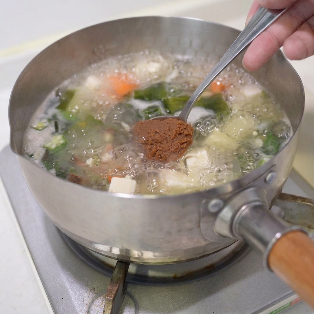 Une cuillère de pâte miso brune plongée dans la soupe de légumes en ébullition.