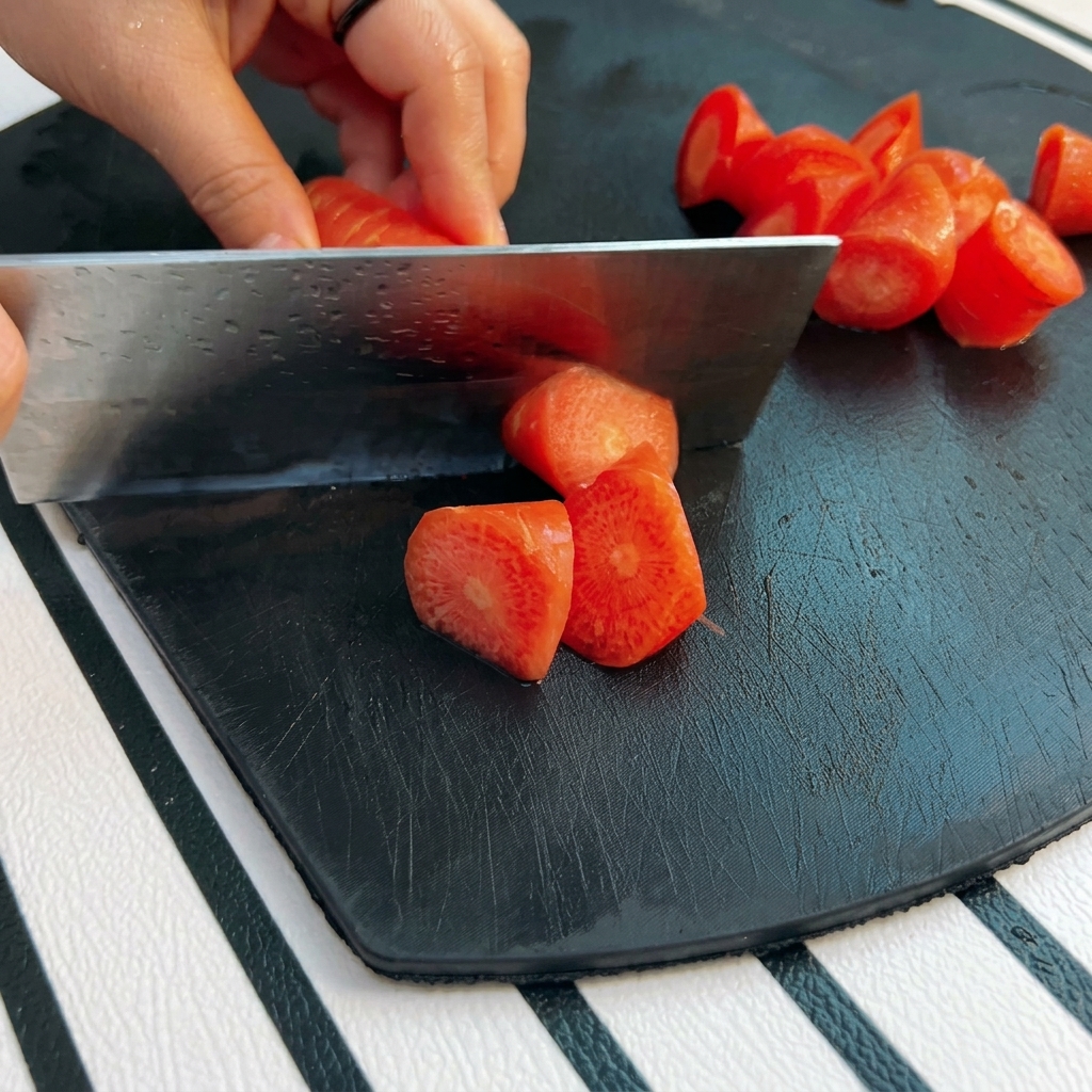 Une main utilisant un grand couteau pour couper une carotte orange vif en morceaux biseautés sur une planche noire.
